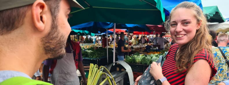 Mirela Letailleur The Travel Bunny et Mathieu au marché de Saint-Paul à La Réunion entourés d’étals de fruits et légumes.