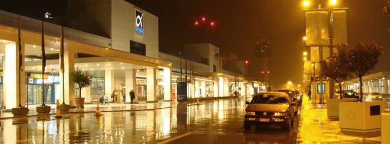 aeroport athenes transport nuit taxi bus 24h Extérieur de l’aéroport international d’Athènes de nuit avec taxis stationnés sous la pluie et terminal éclairé pour transport aéroport Athènes 24h.