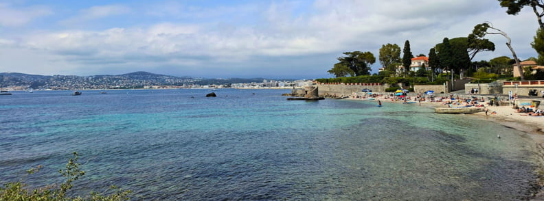 Plage du Port Mallet au Cap d’Antibes avec eau turquoise peu profonde, rivage paisible et vue sur le Golfe Juan à proximité de Posidonia Antibes.