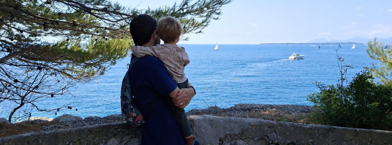 Parent tenant un jeune enfant devant un ancien poste d’observation de la Batterie du Graillon à Posidonia Antibes, avec vue sur la mer et les voiliers du Cap d’Antibes.