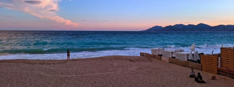 Plage de Cannes en septembre au coucher du soleil, avec la Méditerranée, des transats empilés et les reliefs de l’Estérel au loin.
