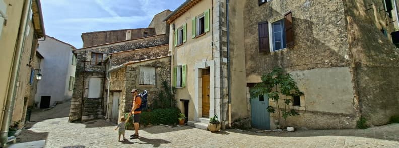 Père et jeune enfant marchant dans les anciennes rues pavées de Mons dans le Var, entre maisons en pierre et façades aux volets colorés.