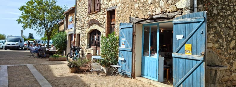 Petit café aux volets bleus dans le centre ancien de Mons dans le Var, avec terrasse sur une rue pavée du village.