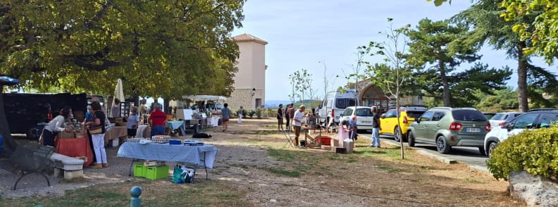 Petit marché installé sur la place Saint-Sébastien à Mons dans le Var, avec stands de produits locaux, visiteurs et voitures garées sous les arbres.