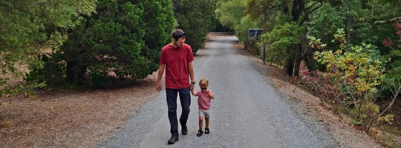 Un père et son jeune enfant marchent main dans la main sur une allée goudronnée du Parc Aurélien à Fréjus, entourée d’arbres et de végétation méditerranéenne.