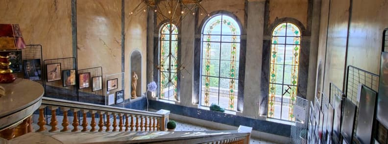 Grand escalier en pierre de la Villa Aurélienne à Fréjus, bordé de balustrades sculptées et de trois hautes fenêtres cintrées à vitraux floraux, avec des photographies exposées sur les murs.