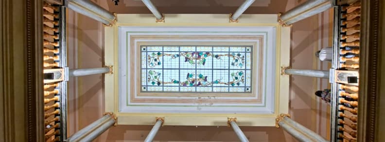 Vue depuis le bas de l’atrium de la Villa Aurélienne à Fréjus, avec une verrière rectangulaire à vitraux floraux, des colonnes claires et des moulures décoratives.