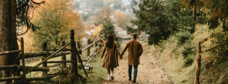 Cuplu îmbrăcat în haine de toamnă plimbându-se pe o alee rurală printre copaci colorați, imagine perfectă pentru escapade romantice de toamnă și călătorii lente în natură.