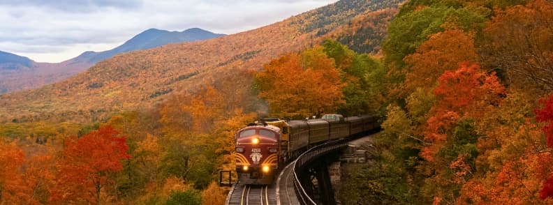 Tren panoramic traversând munți acoperiți de păduri arămii, perfect pentru excursii cu trenul toamna și călătorii lente prin peisaje de toamnă din Europa sau SUA.