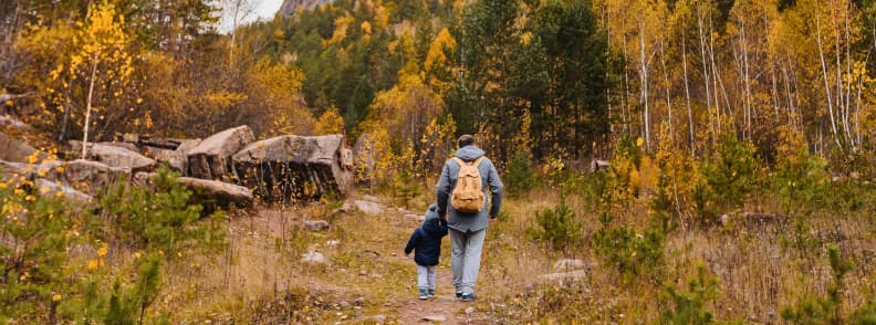 Vacante de toamna pentru familii drumetii si natura colorata Părinte și copil mergând pe o potecă prin pădurea de toamnă, înconjurați de copaci galbeni și roșcați, simbol al unei vacanțe de toamnă pentru familii în natură