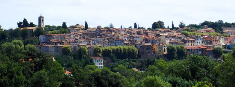 Vedere panoramică asupra satului Bagnols-en-Forêt din Provence, cu biserica Saint-Antonin, case tradiționale cu acoperișuri de țiglă roșie și dealuri împădurite.