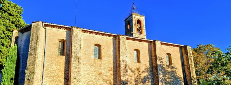 Biserica Saint-Antonin din Bagnols-en-Forêt, Var, cu fațadă din piatră și tencuială ocru, ferestre arcuite și clopotniță sub cerul albastru.