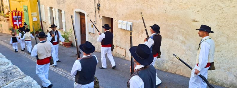 Bravadeurs în costume tradiționale provensale, cu pantaloni albi, veste, eșarfe roșii și muschete, defilând pe o stradă din Bagnols-en-Forêt în spatele unui steag ceremonial.