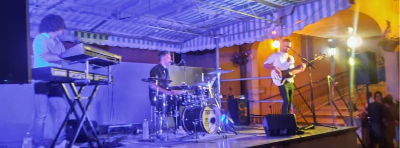 Concert de vară în Bagnols-en-Forêt, cu trei muzicieni cântând la clape, tobe și chitară electrică pe o scenă iluminată în albastru și violet, în timpul unei sărbători locale.