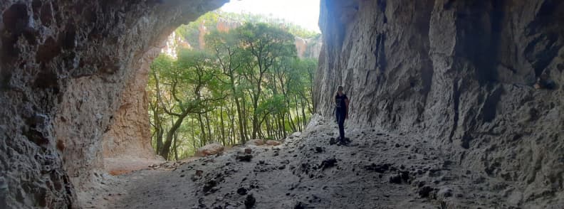 Drumeție la Grotte du Muéron din Bagnols-en-Forêt, cu o persoană lângă peretele stâncos al peșterii, sub o boltă naturală deschisă spre pădure.