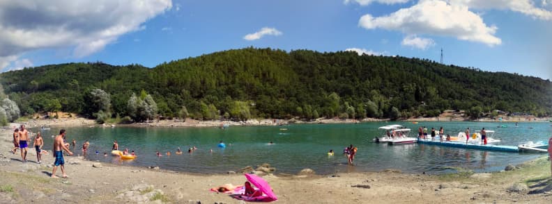 Plajă pe malul Lacului Saint-Cassien din departamentul Var, cu familii care înoată în apa turcoaz, copii pe mal, bărci și o platformă plutitoare lângă dealurile împădurite.