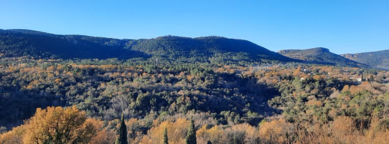 Panorama dealurilor împădurite din jurul satului Bagnols-en-Forêt, în Var, cu arbori în nuanțe aurii de toamnă, chiparoși și creste montane sub cerul senin.