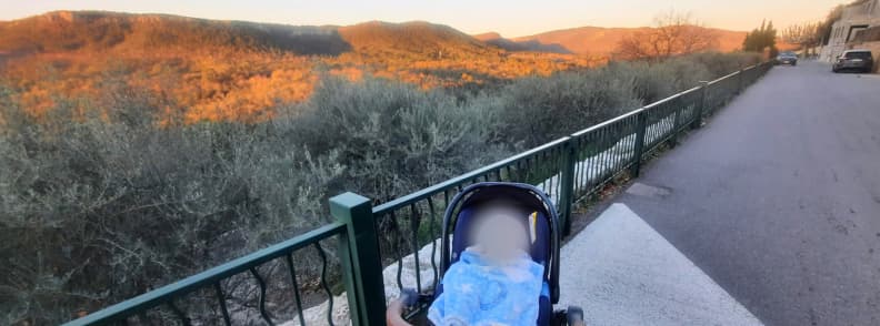 Copil într-un cărucior pe Boulevard du Rayol din Bagnols-en-Forêt, lângă o balustradă verde, cu vedere spre dealurile împădurite din Var luminate de apus.