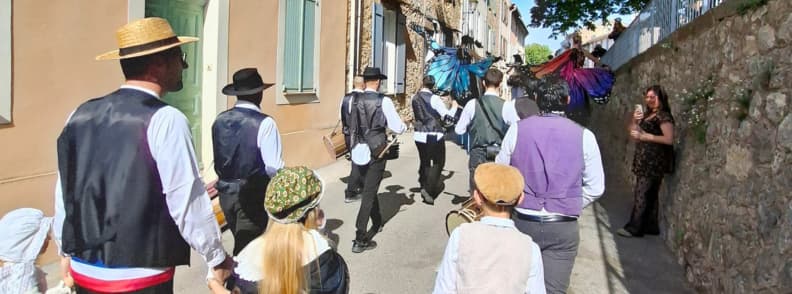 Procesiune festivă pe o stradă din Bagnols-en-Forêt, cu participanți în costume tradiționale provensale, pălării, veste și eșarfe, însoțiți de personaje cu aripi colorate.