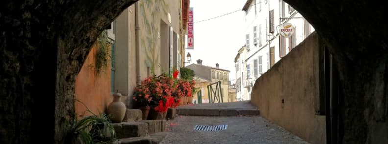 Pasaj boltit din piatră pe Rue de la Voûte din Bagnols-en-Forêt, cu trepte pavate, ghivece cu flori roșii și case tradiționale provensale.