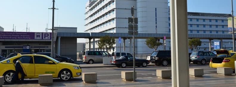 Taxiuri galbene si masini parcate in fata aeroportului Atena langa accesul la trenul suburban Proastiakos