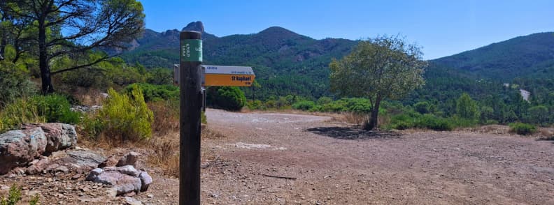 traseu col de belle barbe ravin du gratadis esterel saint raphael Indicator de traseu la Col de Belle Barbe, punct de plecare pentru drumeția Ravin du Gratadis în masivul Estérel, cu dealuri împădurite pe fundal.