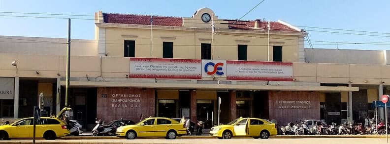 Fațada principală a gării centrale Larissa din Atena cu taxiuri galbene parcate în față și semnul OSE al organizației feroviare elene