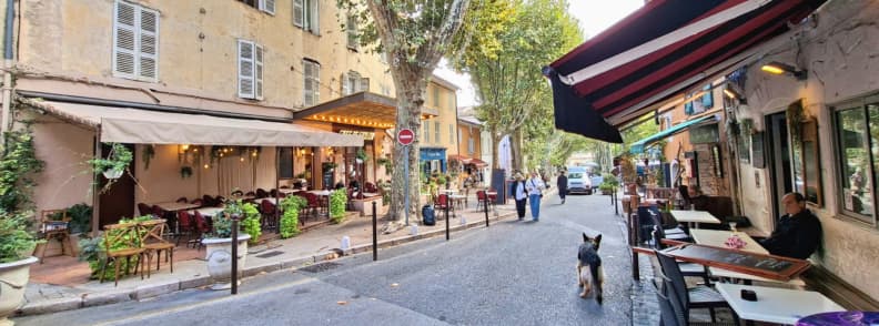 Stradă animată din Cotignac Provence Verte cu terasele Bar Le Marigny și Café de l’Union, decorate cu lumini și plante sub platanii satului
