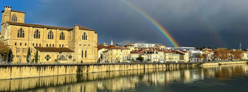 Vedere spre Biserica Saint Barnard si casele istorice din Romans sur Isere cu un curcubeu pe cer si reflexii pe raul Isere