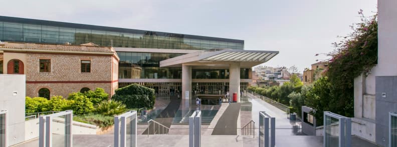 Muzee din Atena muzeul Acropole intrare moderna Intrarea principală în muzeul Acropole din Atena, cu arhitectură modernă, rampă de acces și clădiri istorice în jur.