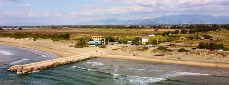Vedere aeriană asupra plajei cu nisip din Anzio, cu dig, valuri, dune joase și munți în depărtare