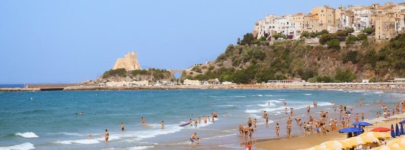 Plajă animată din Gaeta, cu turiști, umbrele, apă albastră și centrul istoric ridicat pe faleză