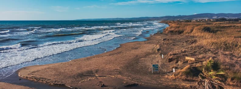 Plajă sălbatică din Ladispoli, cu nisip închis la culoare, valuri, dune joase și vegetație uscată pe coasta Lazio