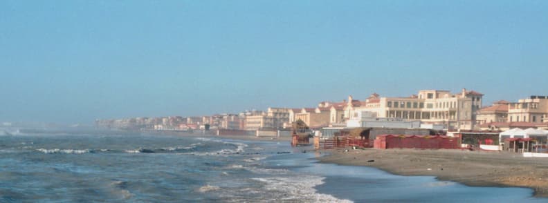 Plaja Lido di Ostia în timpul iernii, cu valuri, nisip aproape gol și clădiri istorice de-a lungul falezei