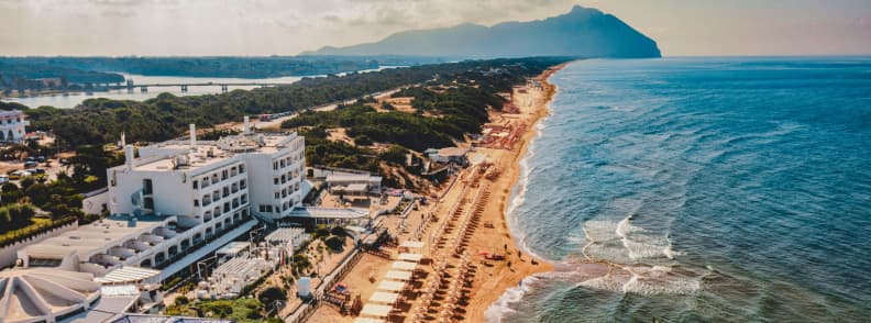Vedere aeriană asupra plajei Sabaudia, cu șiruri de umbrele, hoteluri pe litoral, Marea Tireniană și Monte Circeo în depărtare