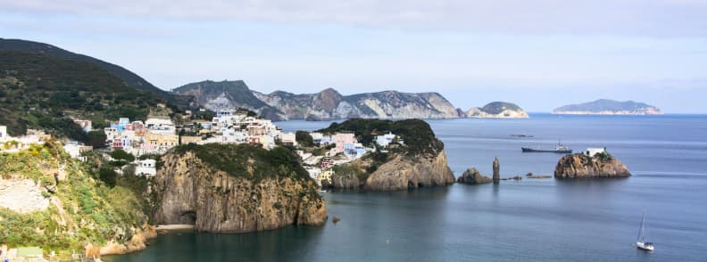 Panoramă asupra insulei Ponza, cu faleze vulcanice, case albe, golfuri cu apă limpede și insulele Pontine în fundal