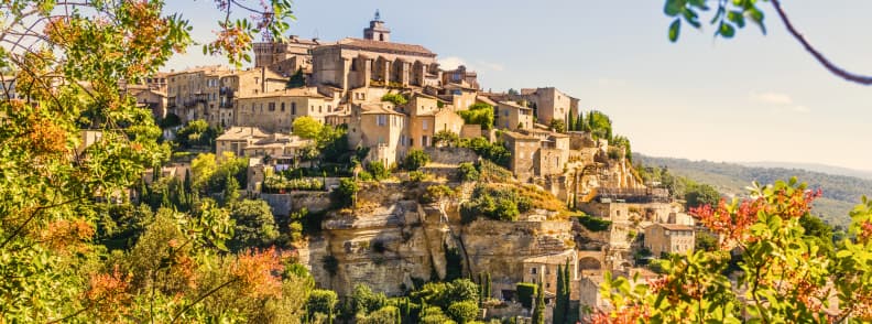 Gordes Luberon Provence Toamna Culori de Toamna Gordes în Luberon toamna, cu case din piatră ridicate pe stâncă, vegetație în nuanțe aurii și roșiatice și dealurile Provence în fundal.