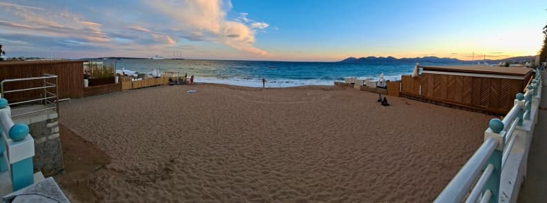 Plajă aproape goală din Cannes toamna, la apus, cu nisip, șezlonguri strânse, Marea Mediterană și dealurile Estérel la orizont.