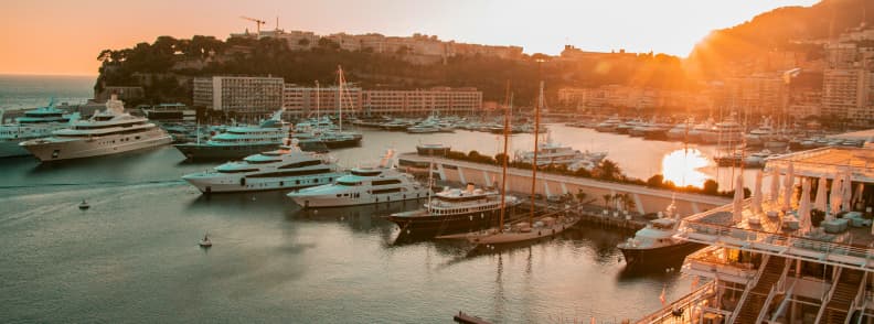 Port Hercule din Monaco la apus toamna, cu superiahturi acostate, Monte Carlo pe versant și lumină aurie peste port.
