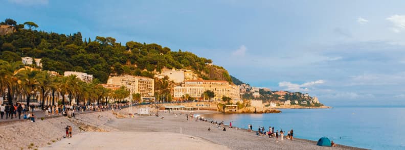 Toamna pe Coasta de Azur. Plajă cu pietricele din Nisa toamna, cu Promenade des Anglais, palmieri, oameni pe mal și Marea Mediterană calmă.