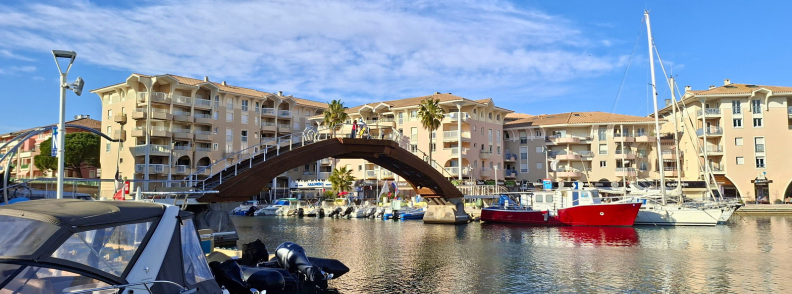 Port Frejus cazare marina restaurante si plimbari Port-Fréjus cu ambarcațiuni acostate, pod pietonal curbat, apartamente pe mal și palmieri în apropierea marinei.