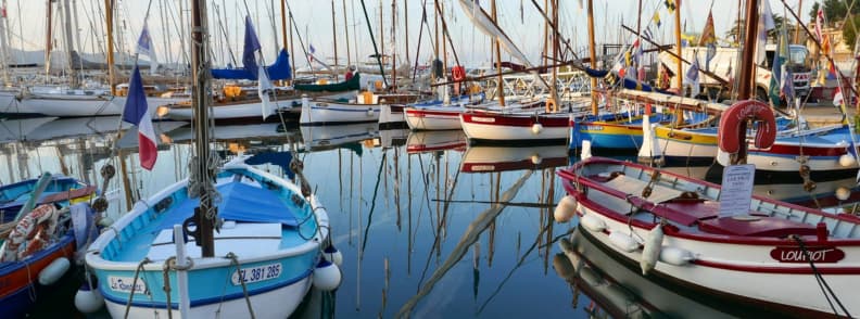 Veliere tradiționale și ambarcațiuni colorate acostate într-un port de pe Riviera Franceză, cu catarge reflectate în apa calmă.