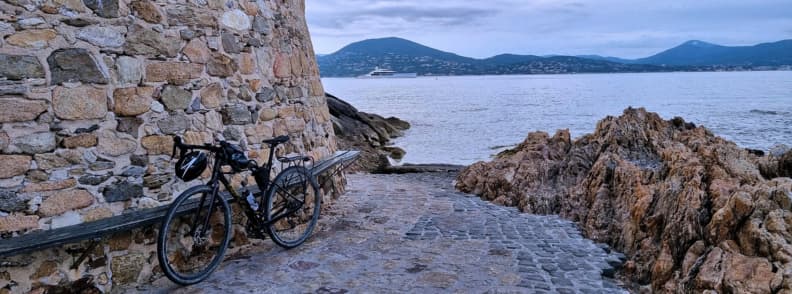 Bicicletă sprijinită de un zid de piatră pe coasta din Saint-Tropez, cu stânci, Marea Mediterană și dealurile Golfului Saint-Tropez în fundal.