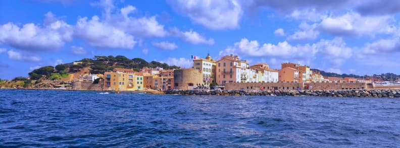Saint-Tropez văzut de pe mare toamna, cu clădiri pastelate, ziduri de piatră, portul și dealurile verzi de pe Coasta de Azur.