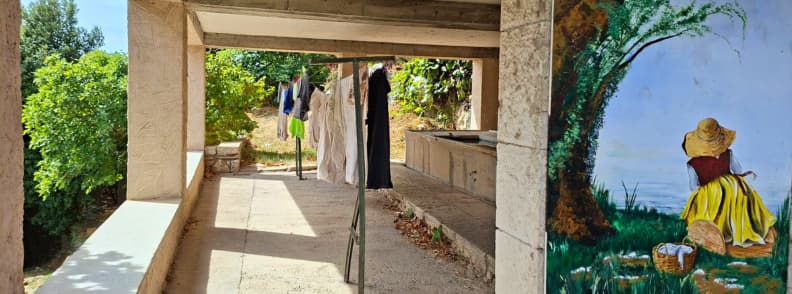 Lavoir istoric Mons Var patrimoniu sat vechi Lavoir acoperit din Mons Var, cu bazine din piatră, rufe întinse și o pictură murală pe peretele vechii spălătorii.