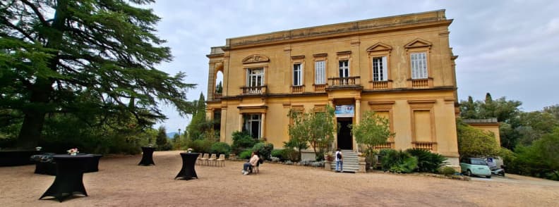Fațada principală a Villa Aurélienne din Fréjus, cu arhitectură de inspirație palladiană, ferestre înalte, balcoane ornamentale și terasă cu pietriș, înconjurată de copaci.