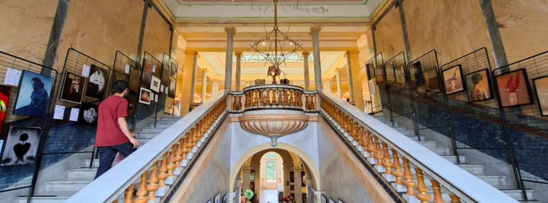 Scara monumentală dublă din Villa Aurélienne din Fréjus, cu balustrade sculptate, fotografii expuse pe pereți și un vizitator care urcă treptele în timpul festivalului de fotografie suprarealistă.