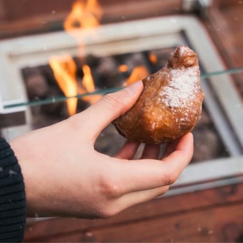 Jordaan food tour Dutch Street Food Pastry Amsterdam Snack Jordaan food tour. Hand holding a fried Dutch pastry dusted with sugar near an outdoor fire in Amsterdam