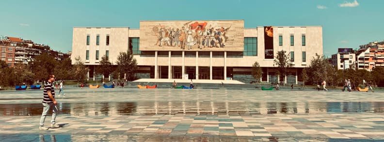 Local Tips for Tirana Safety Language and Connectivity Guide The National History Museum faces Skanderbeg Square in central Tirana, with pedestrians crossing the wide open plaza on a sunny day.