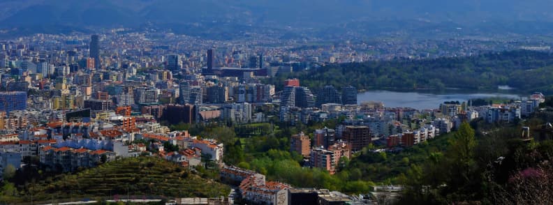 Panoramic view of Tirana Albania, with the city center, Grand Park lake, surrounding neighborhoods, and mountains in the distance.